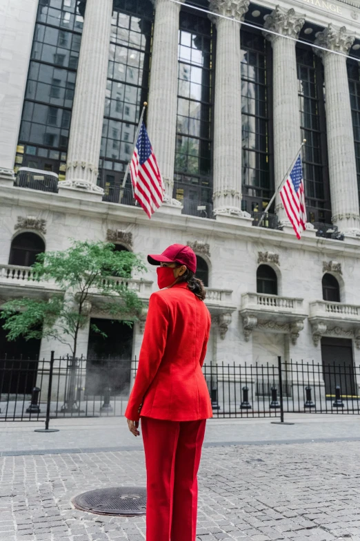 a woman in a red suit stands in front of the new york stock exchange, by Nina Hamnett, trending on unsplash, red ascot and a field cap, wearing track and field suit, facing away from camera, environmental shot
