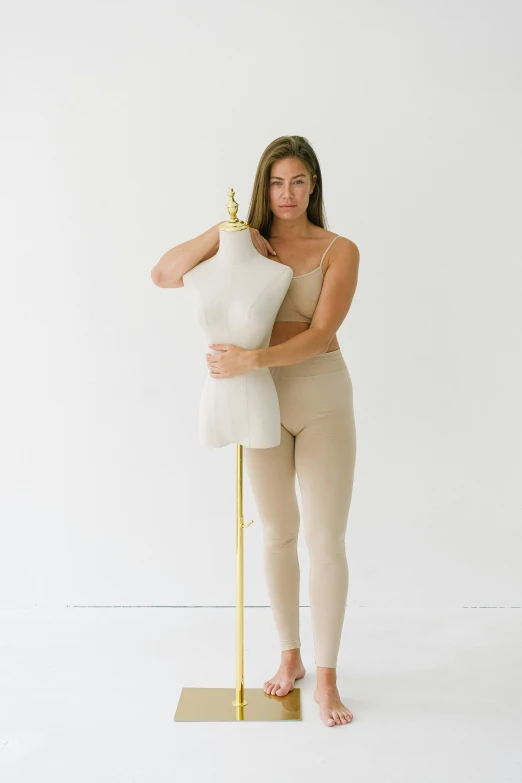 a woman standing next to a mannequin on a stand, featured on instagram, renaissance, wearing leotard, plain background, beige and gold tones, devon cady-lee