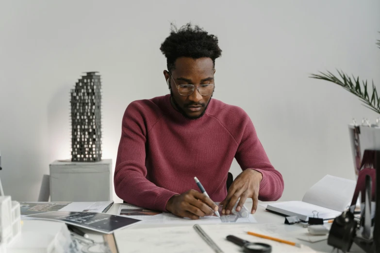 a man sitting at a table writing on a piece of paper, a drawing, inspired by Afewerk Tekle, pexels contest winner, architect studio, he is wearing a brown sweater, avatar image, virgil abloh