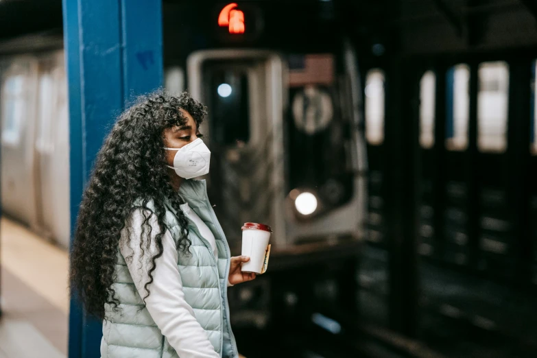 a woman standing on a train platform with a cup of coffee, trending on pexels, graffiti, people are wearing masks, african american woman, ny, drinking cough syrup
