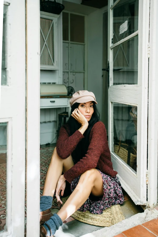 a woman sitting in a doorway talking on a cell phone, a picture, by Lucia Peka, trending on pexels, maroon hat, korean women's fashion model, wearing casual sweater, short in stature