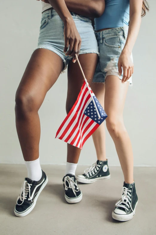 two women standing next to each other holding an american flag, a portrait, trending on unsplash, bra and shorts streetwear, shows a leg, wearing white sneakers, alexis franklin