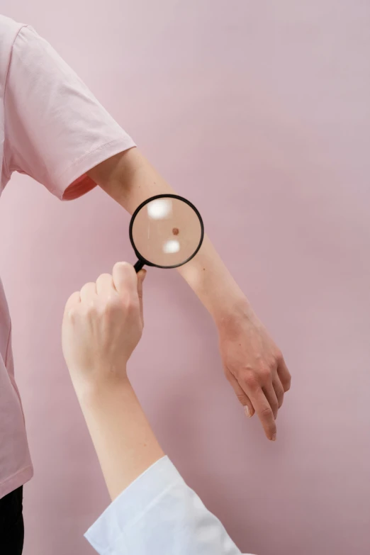 a person holding a magnifying glass over another person's arm, a tattoo, pastel pink skin tone, mole on cheek, skincare, poorly lit