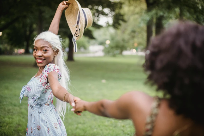 a woman holding a hat on top of a lush green field, by Lily Delissa Joseph, pexels contest winner, dancing with each other, black young woman, girl with white hair, flirting smiling