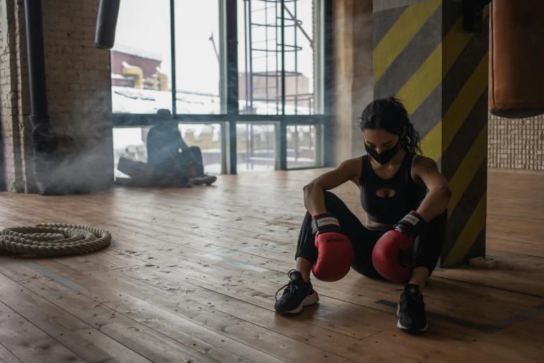 a woman sitting on the floor in front of a punching bag, by Emma Andijewska, pexels contest winner, mask off, athletic crossfit build, manuka, in a massive cavernous iron city