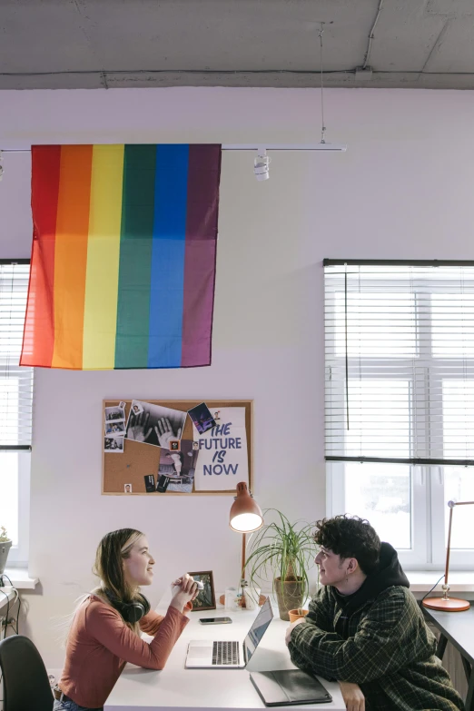 a couple of people sitting at a table with laptops, by Harriet Zeitlin, trending on unsplash, lgbt flag, sitting in dean's office, hung above the door, low ultrawide shot