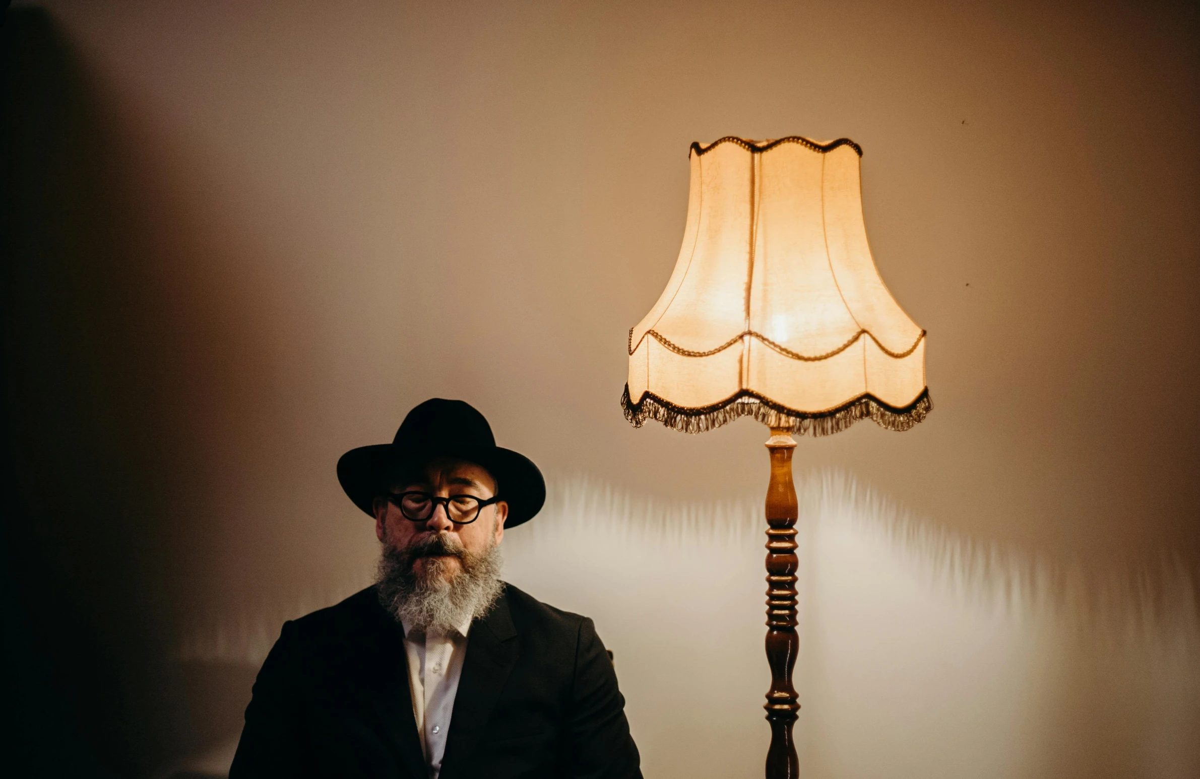 a man in a suit and hat sitting in front of a lamp, an album cover, by Will Ellis, unsplash, long grey beard, jewish young man with glasses, floor lamps, looking directly at the viewer