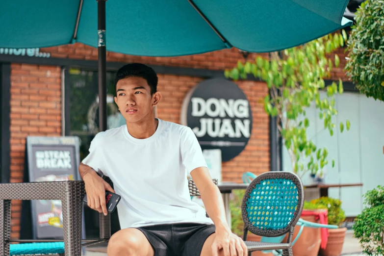 a man sitting on a chair under an umbrella, inspired by Dong Yuan, instagram, dau-al-set, tan skin a tee shirt and shorts, teenager hangout spot, at the counter, johnson ting