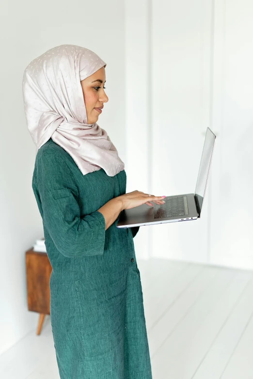 a woman in a green dress holding a laptop, inspired by Maryam Hashemi, hurufiyya, wearing a scarf, grey, bent - over posture, medium height