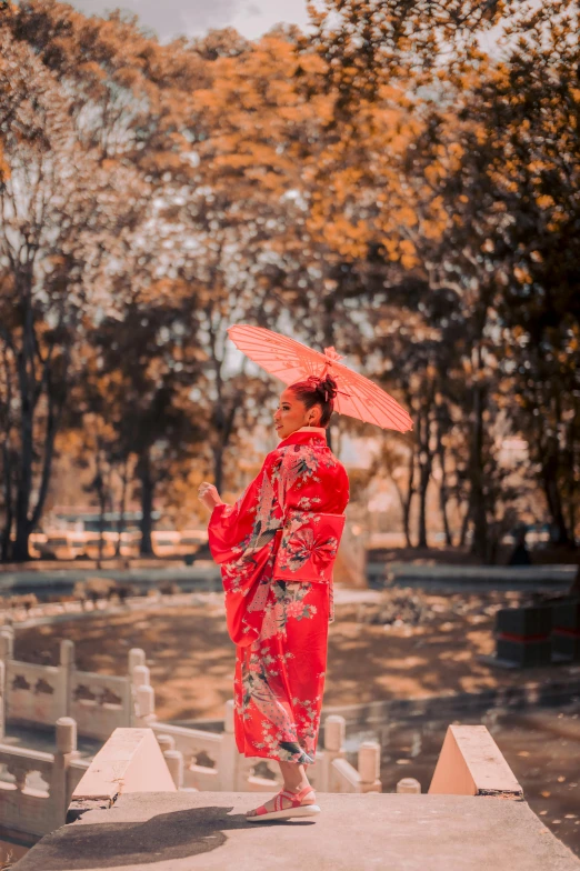 a woman in a red kimono walking across a bridge, a picture, pexels contest winner, standing with a parasol, autumnal empress, sunny day time, posing for a picture