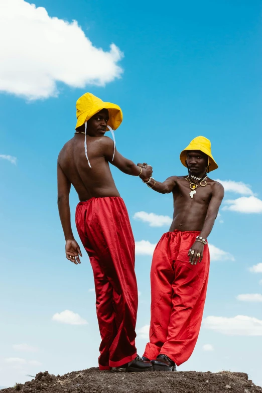 a couple of men standing on top of a hill, an album cover, by Nathalie Rattner, trending on pexels, afrofuturism, wearing red and yellow clothes, red hat, loincloth, virgil abloh