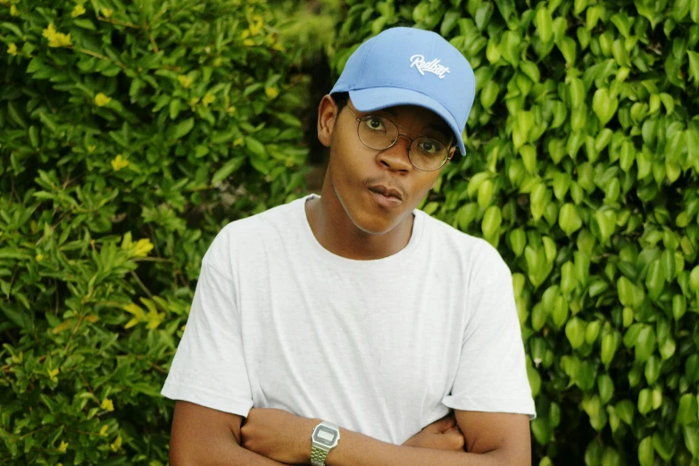 a man standing in front of a bush with his arms crossed, an album cover, by Richmond Barthé, pexels, realism, wearing a baseball cap, smooth blue skin, headshot profile picture, mongezi ncaphayi