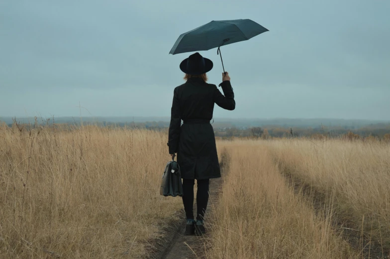 a woman walking down a dirt road holding an umbrella, in a field, wearing black overcoat, instagram picture, prairie