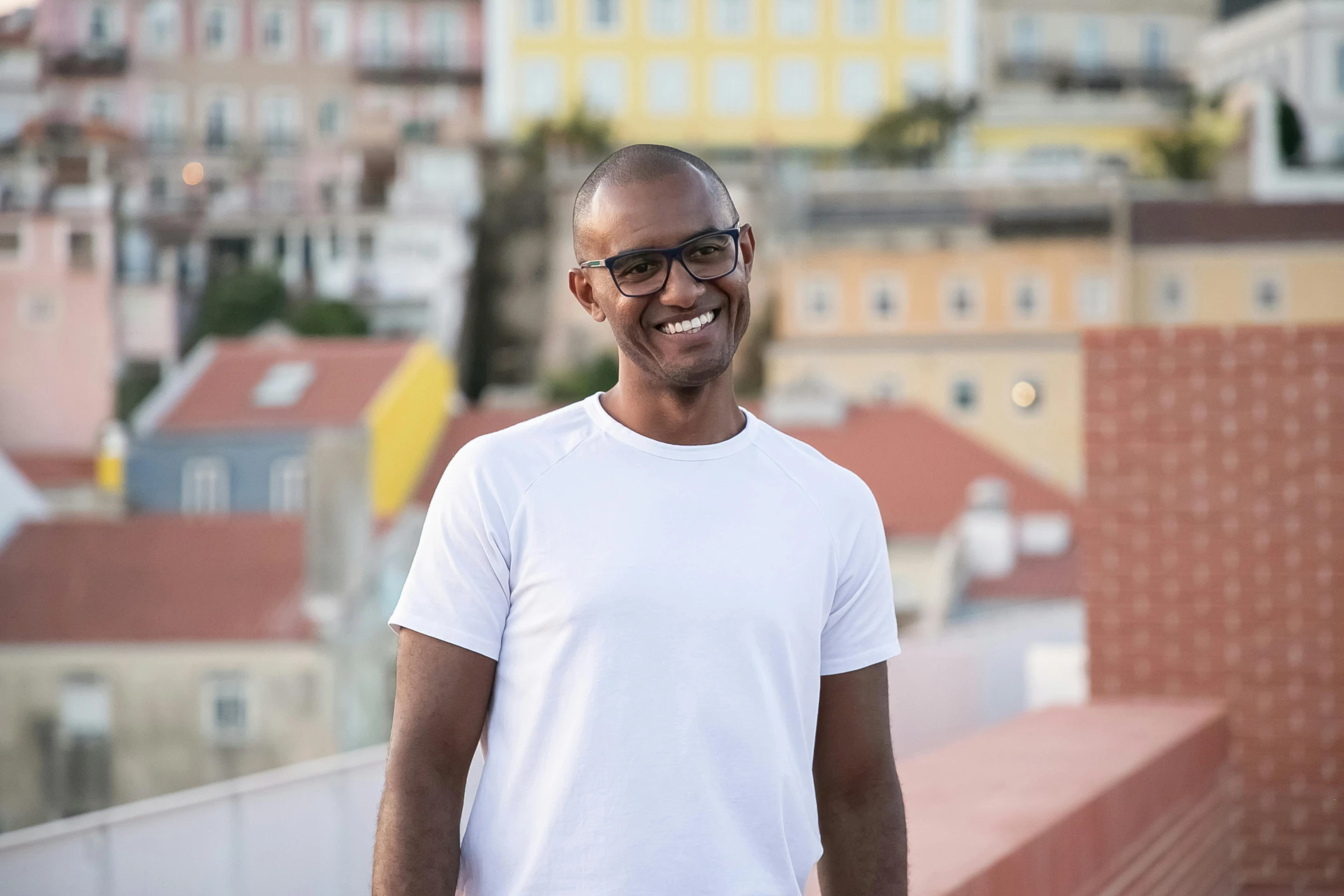 a man standing on top of a roof with buildings in the background, inspired by Francis Souza, happening, smiling at camera, man with glasses, man is with black skin, profile image