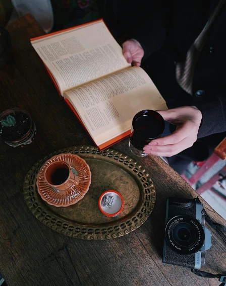 a person sitting at a table with a camera and a book, a still life, pexels contest winner, black and terracotta, carrying a tray, low quality photo, ☕ on the table