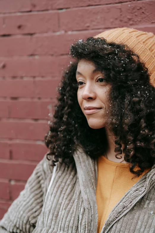 a woman standing in front of a brick wall, a portrait, by Winona Nelson, trending on unsplash, curly black hair, warm muted colors, hat and hoodie, rebecca sugar