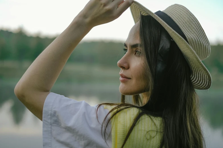 a woman wearing a hat standing next to a body of water, relaxed eyebrows, a woman's profile, trending photo, pondering