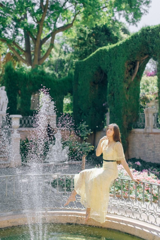 a woman in a yellow dress standing in front of a fountain, lush garden surroundings, belle delphine, splashing, mediterranean features