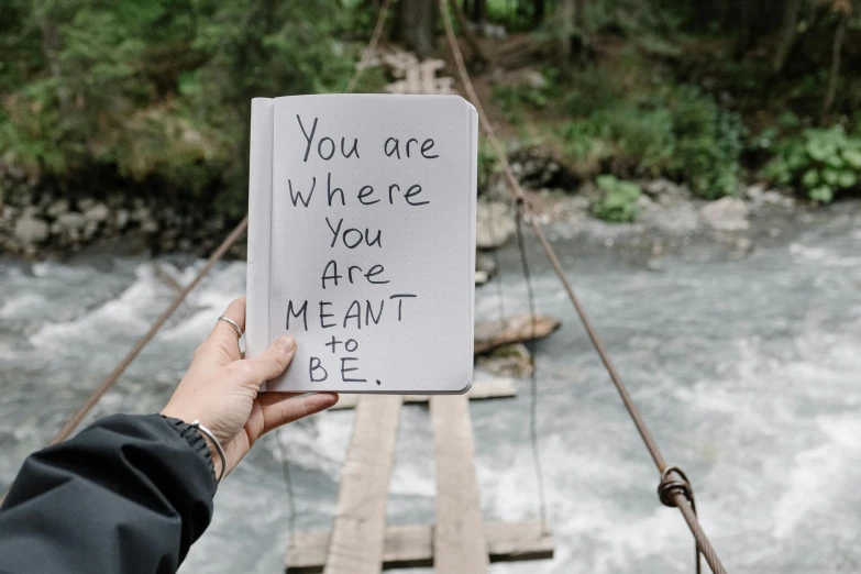 a person holding a sign that says you are where you are meant to be, by Caroline Mytinger, pexels contest winner, rope bridge, notebook, yand.re, heath clifford