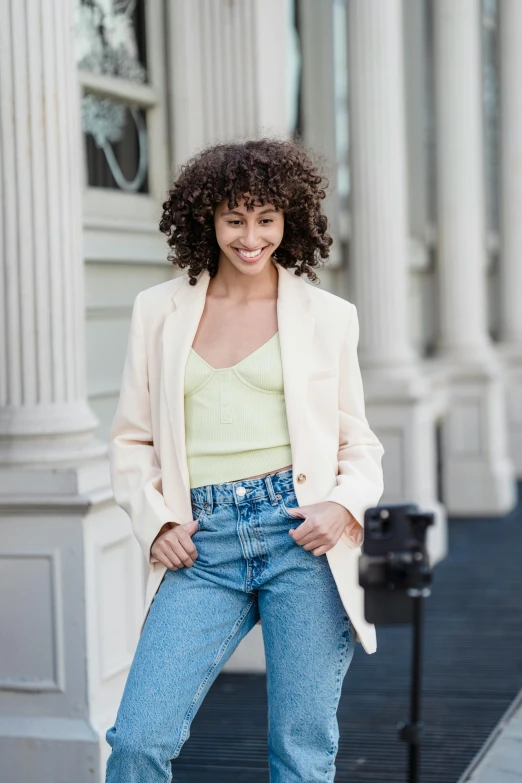 a woman posing for a picture in front of a building, wearing jeans, light yellow, with a curly perm, wearing a blazer