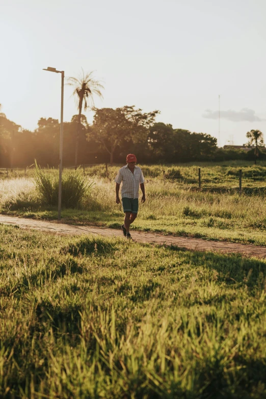 a man walking down a dirt road next to a lush green field, a portrait, unsplash, in sao paulo, evening sun, suburb, people running