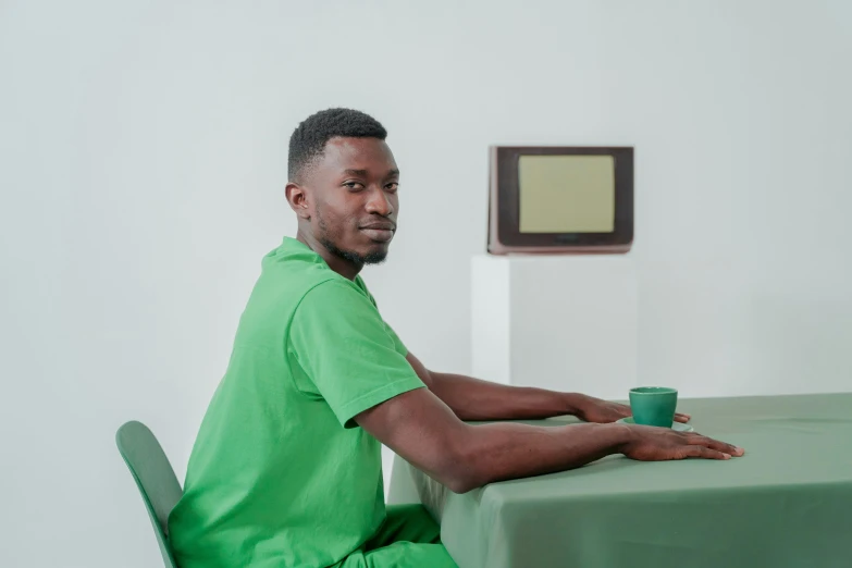 a man sitting at a table with a cup of coffee, green colored skin, david uzochukwu, healthcare worker, waiting room