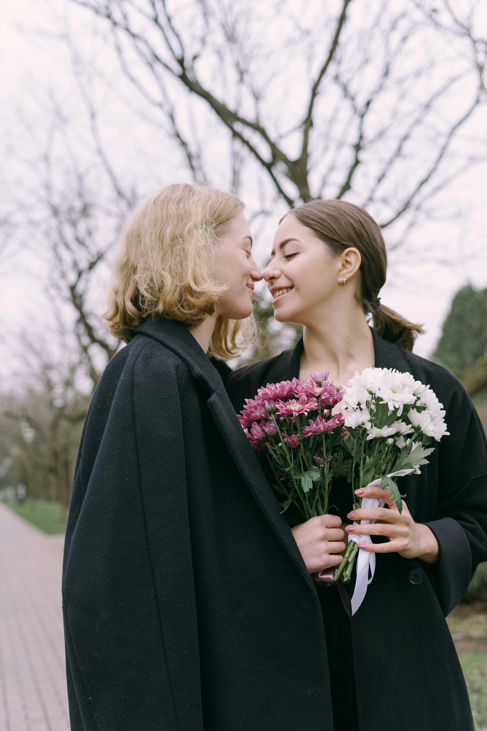 a couple of women standing next to each other, trending on unsplash, romanticism, carrying flowers, wearing black coat, lesbian kiss, orthodox