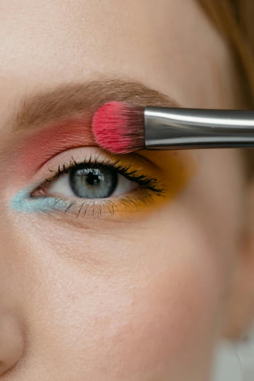 a close up of a person with a brush on their eye, wearing eye shadow, red and blue eyes, multi - coloured, easy to use