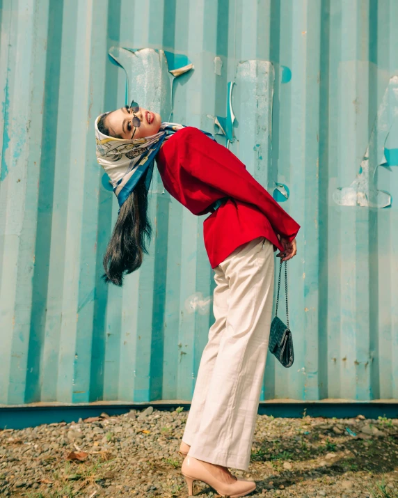 a woman standing in front of a blue wall, trending on pexels, hurufiyya, red sweater and gray pants, white hijab, bella poarch, next to a red barn