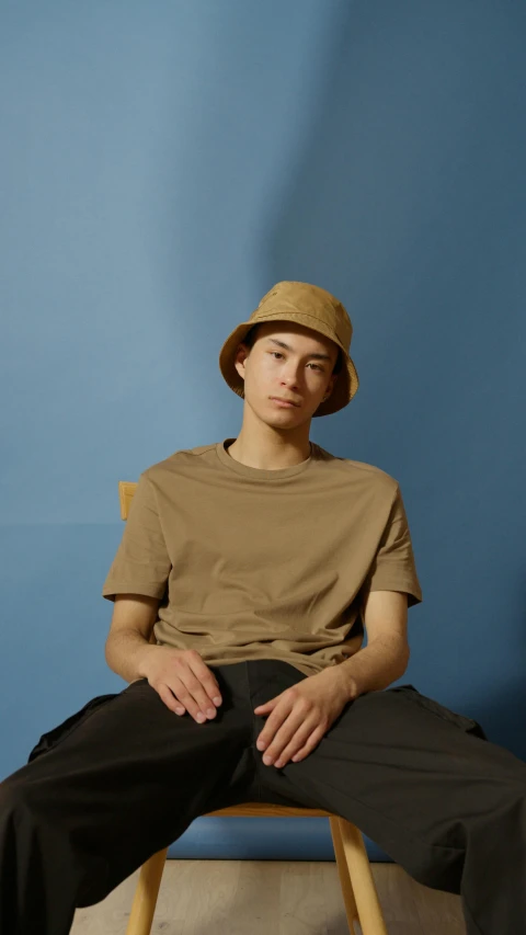 a man sitting on top of a wooden chair, an album cover, inspired by Russell Dongjun Lu, pexels contest winner, realism, bucket hat, light brown, wearing a t-shirt, default pose neutral expression