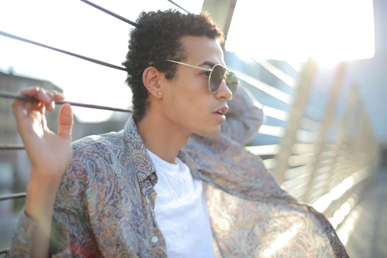 a man wearing sunglasses leaning against a fence, an album cover, pexels, hurufiyya, light-brown skin, mixed race, androgynous person, patterned clothing