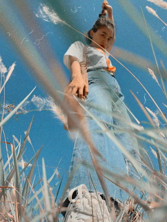 a woman standing on top of a grass covered field, an album cover, inspired by Ren Hang, trending on unsplash, magic realism, wearing a light blue shirt, wearing farm clothes, ☁🌪🌙👩🏾, orange grass