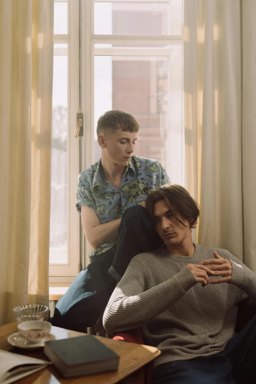 two men sitting on a couch in front of a window, an album cover, trending on pexels, androgynous person, two men hugging, alexey gurylev, ( ( theatrical ) )