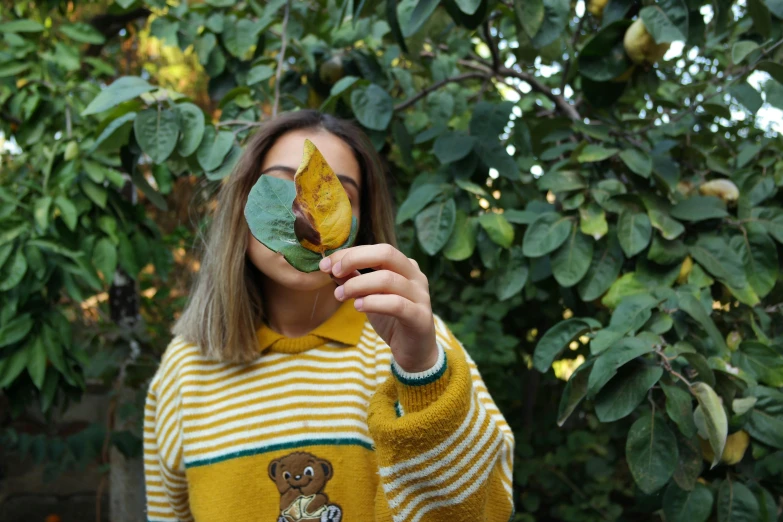 a woman holding a leaf in front of her face, inspired by Elsa Bleda, pexels contest winner, striped sweater, wearing yellow croptop, 🍂 cute, taken on iphone 14 pro