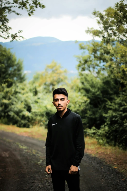 a man standing in the middle of a dirt road, wearing a black hoodie, asher duran, in front of a forest background, ((portrait))