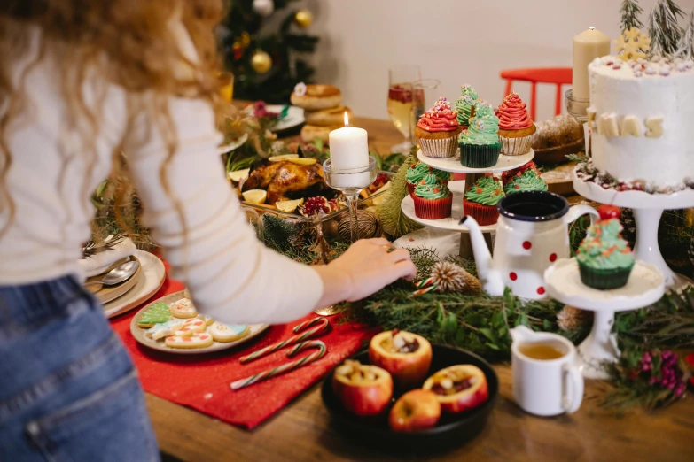 a woman standing in front of a table filled with cupcakes, by Julia Pishtar, pexels, christmas, vegetables on table and candle, avatar image, table in front with a cup