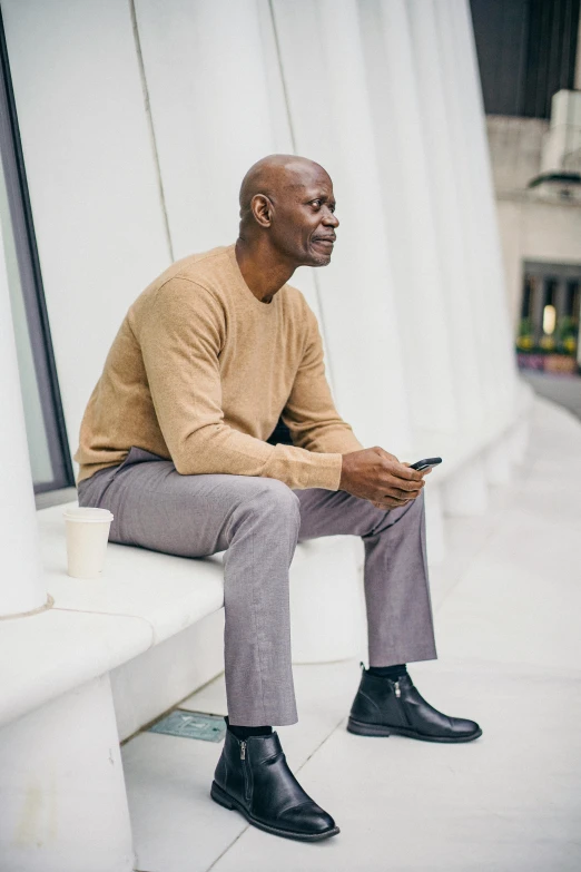 a man sitting on a bench using a cell phone, brown sweater, lance reddick, tailored clothing, curated collections