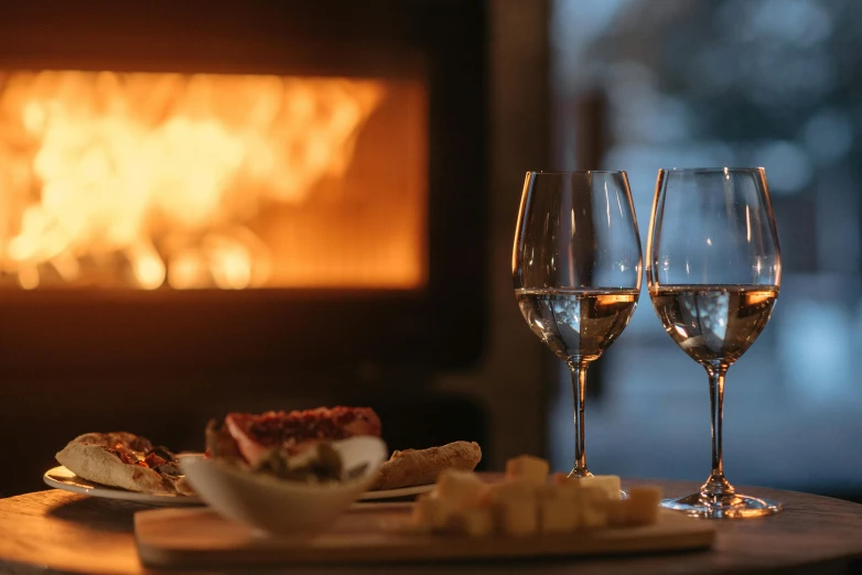 a couple of wine glasses sitting on top of a table, by Carey Morris, pexels contest winner, cosy fireplace, eating a cheese platter, winter setting, local close up