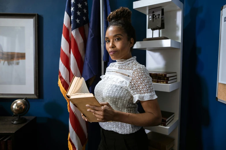 a woman holding a book in front of an american flag, standing in a dimly lit room, mixed-race woman, defense attorney, the librarian