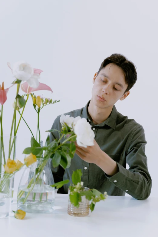 a man sitting at a table with flowers in vases, an album cover, inspired by Fei Danxu, romanticism, low quality footage, pose 4 of 1 6, picking up a flower, a portrait of issey miyake