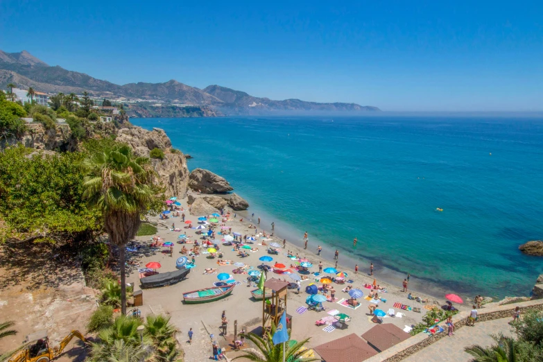 a beach filled with lots of people next to the ocean, in spain, monkey island, cliffside town, clear blue water