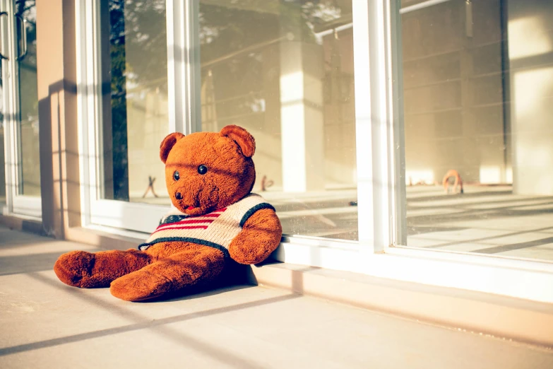 a teddy bear sitting in front of a window, a cartoon, by Julia Pishtar, pexels contest winner, harsh sunlight, schools, william eggleston style, sitting on the ground