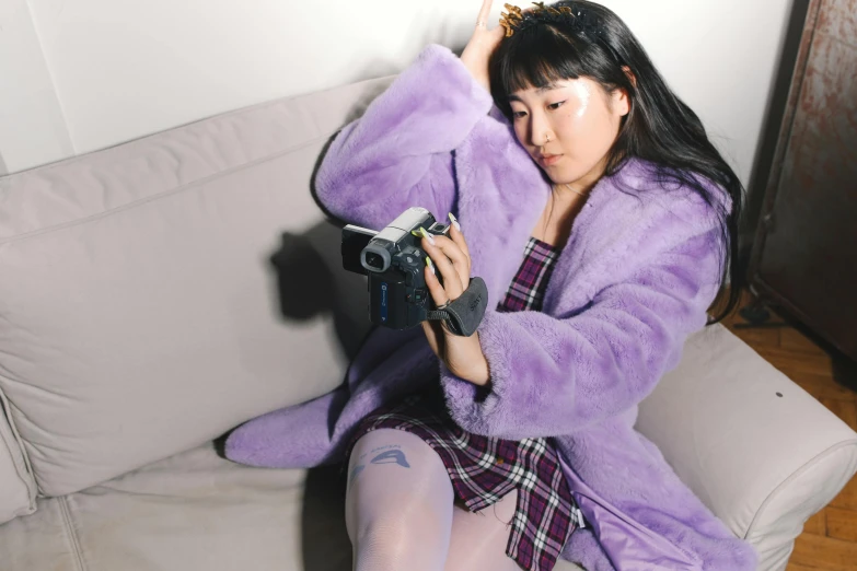 a woman sitting on a couch holding a camera, an album cover, inspired by Yuko Tatsushima, trending on pexels, wearing a purple detailed coat, cosplay photo, fluffy full of light, in an action pose
