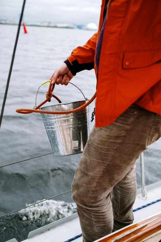 a person standing on a boat holding a bucket, up close, grey orange, hydration, sailor clothing