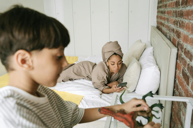 a woman sitting on top of a bed next to a boy, trending on pexels, visual art, playing games, phone photo, green and brown clothes, modest