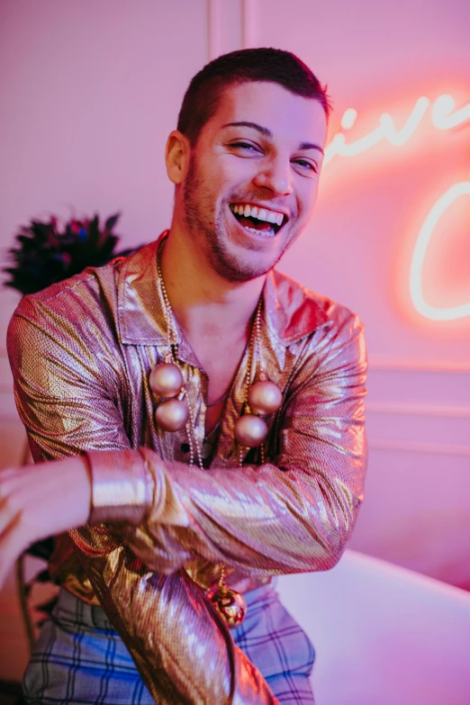 a man sitting on a couch in front of a neon sign, an album cover, by Julia Pishtar, trending on pexels, wearing shiny gold catsuit, mutahar laughing, young greek man, full of sand and glitter