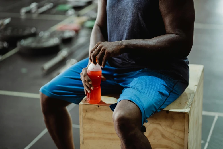 a man sitting on top of a wooden box holding a drink, local gym, profile image, wearing red shorts, spilling juice