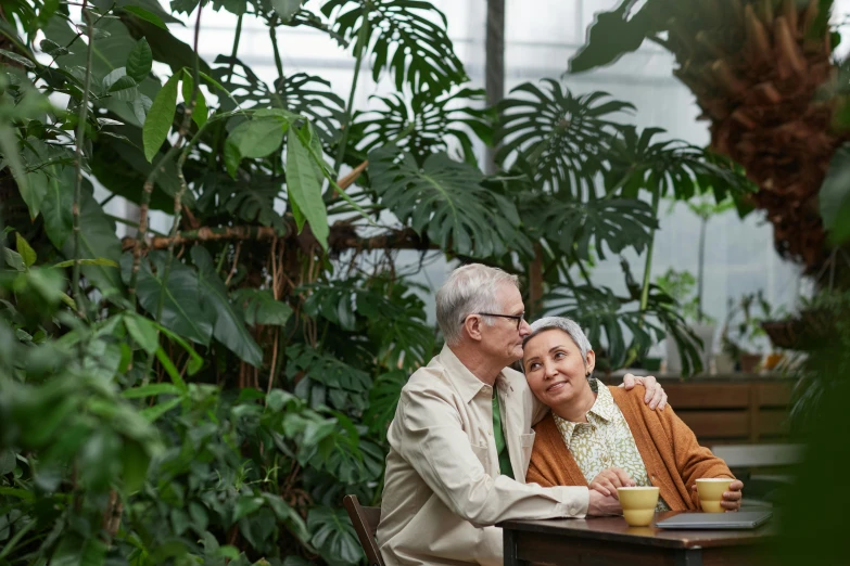 a man and woman sitting at a table in a greenhouse, pexels contest winner, two old people, lush foliage, hugging, sheltering under a leaf