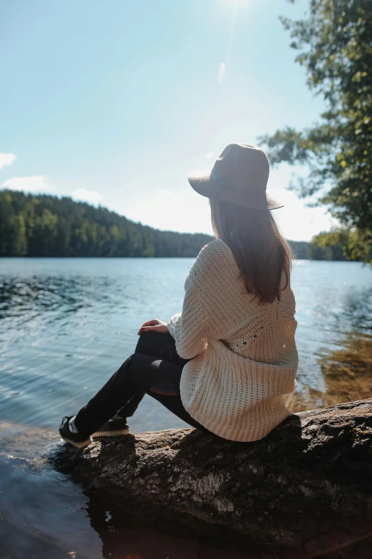 a woman sitting on a rock in front of a lake, wearing a white sweater, wearing a travel hat, pondering, 2019 trending photo