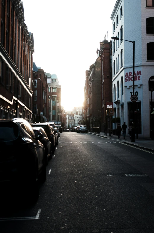 a street lined with parked cars next to tall buildings, a picture, unsplash, winter sun, london, late summer evening, gothic city streets behind her